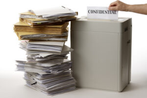 A large stack of disorganized papers and folders is beside a shredder. A hand inserts a sheet labeled "CONFIDENTIAL" into the shredder.
